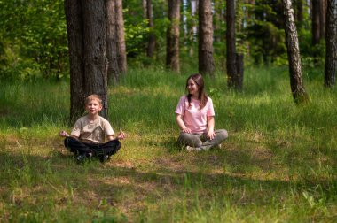 Anne ve oğlu orman çimlerinde bağdaş kurmuş, uyum içinde meditasyon yapıyorlar.