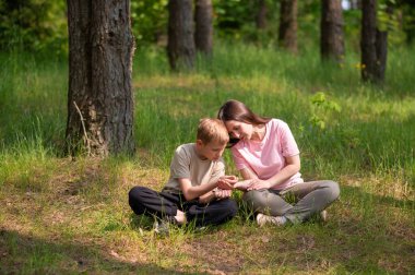 Anne ve oğlu bir ormanda yan yana oturmuş avuçlarındaki bir şeye bakıyorlar.