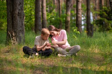 Anne ve oğlu bir ormanda yan yana oturmuş avuçlarındaki bir şeye bakıyorlar.