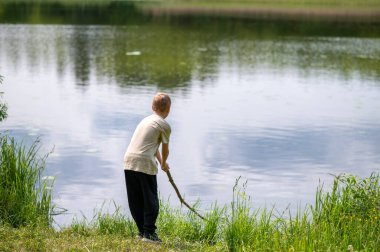 Çocuk uzun bir sopayla gölde duruyor, doğada bir yaz günü suyun yanında oynuyor.