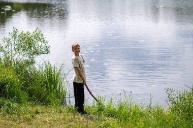 Çocuk uzun bir sopayla gölde duruyor, doğada bir yaz günü suyun yanında oynuyor.
