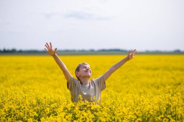 Kollarını açmış neşeli bir çocuk mavi gökyüzünün altındaki sarı açan kolza tohumu tarlasında duruyor.