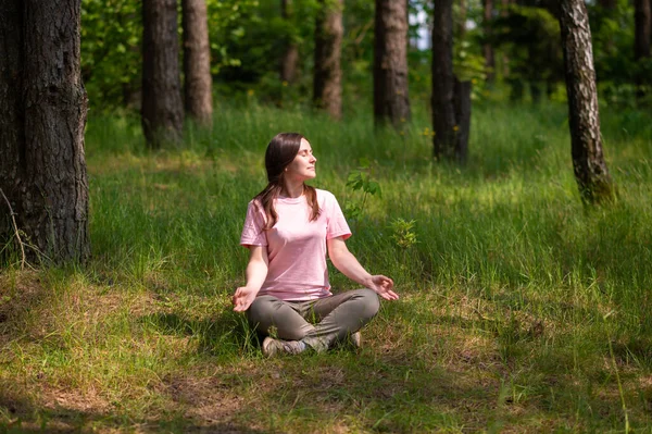 Huzurlu bir ormanda tek başına meditasyon yapan genç bir kadın, yeşil çimlerin üzerinde bağdaş kurarak oturuyor.
