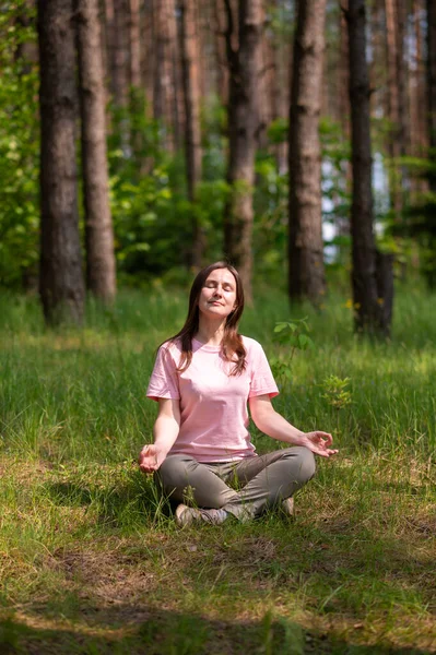 Huzurlu bir ormanda tek başına meditasyon yapan genç bir kadın, yeşil çimlerin üzerinde bağdaş kurarak oturuyor.
