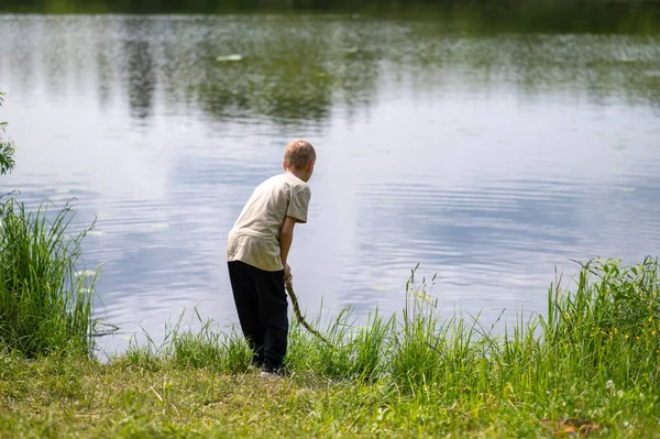 Çocuk uzun bir sopayla gölde duruyor, doğada bir yaz günü suyun yanında oynuyor.