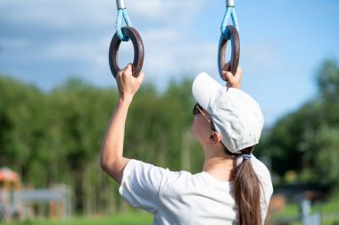 Şapkalı ve güneş gözlüklü genç bir kadın açık hava spor sahasında jimnastik yüzükleri, spor konsepti, güç ve sağlıklı yaşam tarzı eğitimi alıyor.
