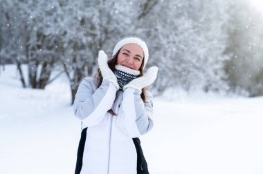 Kar parkında yürürken sıcak beyaz ceket ve eldiven giyen mutlu bir kadın. Sıcak kış yaşam tarzı, olumlu duygular, soğuk hava modası, açık hava eğlencesi ve refah konsepti.