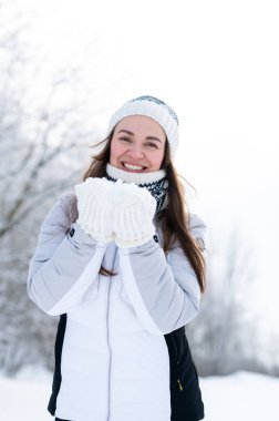 Sıcak kışlık giysiler içinde gülümseyen genç bir kadın soğuk bir parkta karlı bir günün tadını çıkarıyor. Mevsimlik açık hava yaşam tarzı, kış havası, neşe, soğuk hava, doğa, rahatlık ve sıcak kış atmosferi.