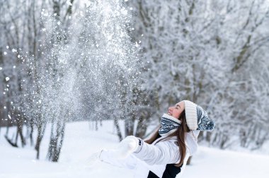 Kış parkında havaya kar atan mutlu genç bir kadın. Eğlenceli kış yaşam tarzı, neşe, soğuk hava, açık hava aktivitesi ve mevsimsel mutluluk.