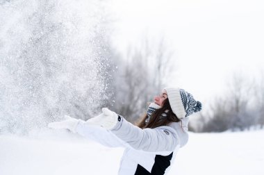 Kış parkında havaya kar atan mutlu genç bir kadın. Eğlenceli kış yaşam tarzı, neşe, soğuk hava, açık hava aktivitesi ve mevsimsel mutluluk.