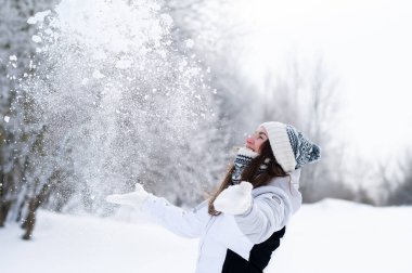 Kış parkında havaya kar atan mutlu genç bir kadın. Eğlenceli kış yaşam tarzı, neşe, soğuk hava, açık hava aktivitesi ve mevsimsel mutluluk.