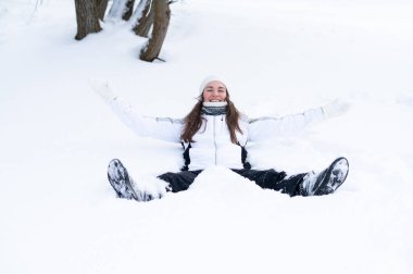 Genç bir kadın taze karda oturuyor, kolları açık, gülümsüyor ve özgürlüğün, sakinliğin ve karlı bir ormanda kış atmosferinin tadını çıkarıyor.
