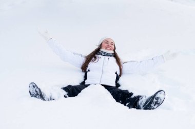 Genç bir kadın taze karda oturuyor, kolları açık, gülümsüyor ve özgürlüğün, sakinliğin ve karlı bir ormanda kış atmosferinin tadını çıkarıyor.