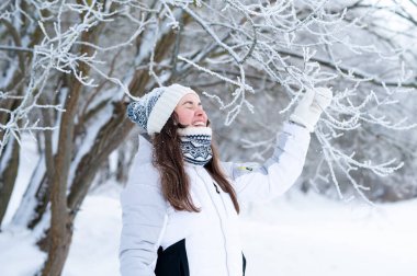 Genç bir kadın kış parkında karlı ağaç dallarına dokunuyor. Sakin kış yaşam tarzı, doğayla bağlantı, soğuk hava ve huzurlu mevsimsel eğlence.