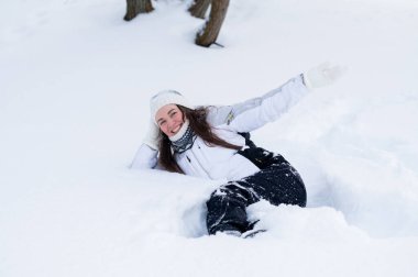 Sıcak kış kıyafetleri içinde gülümseyen genç bir kadın derin taze karda uzanıyor ve karlı bir ormanda huzurlu bir kış gününün tadını çıkarıyor.