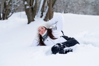 Sıcak kış kıyafetleri içinde gülümseyen genç bir kadın derin taze karda uzanıyor ve karlı bir ormanda huzurlu bir kış gününün tadını çıkarıyor.