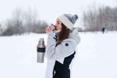 Karlı bir parkta kış yürüyüşü sırasında termostan sıcak çay içen mutlu kadın.