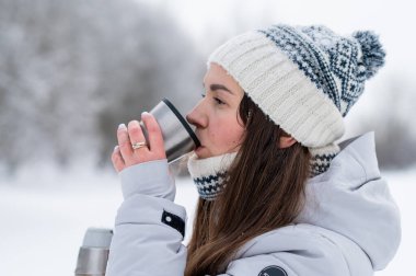 Karlı bir parkta kış yürüyüşü sırasında termostan sıcak çay içen mutlu kadın.