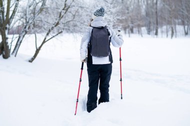 Karlı bir parkta yürüyüş direkleri ile yürüyen bir kadının arka görüntüsü. Kış tatilleri, soğuk sezon sporları, bağımsızlık ve aktif yaşam tarzı konsepti.