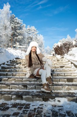 Kürk şapkalı, ceketli ve eldivenli bir kadın kış parkında karla kaplı taş merdivenlerde oturuyor. Rahat bir hava, soğuk güneşli bir gün, mevsimlik moda ve rahat bir yaşam tarzı konsepti.