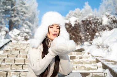 Kürk şapkalı ve ceketli bir kadın karlı merdivenlerde oturuyor ve ellerinden kar üflüyor. Neşeli kış havası, soğuk hava eğlencesi ve mevsimlik açık hava yaşam tarzı portresi.