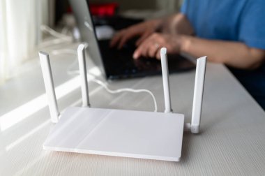 Beyaz ahşap bir masada dört anten bulunan modern bir internet yönlendiricisinin üst görüntüsü. Dişi eller arka planda dizüstü bilgisayarda yazarken görülüyor, uzak iş veya damızlık için yüksek hızlı bağlantı gösteriliyor