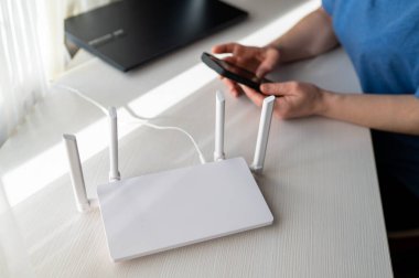 Beyaz bir masada antenleri olan modern kablosuz internet yönlendirici. Arka planda bir kişi kırmızı bir kitap ve dizüstü bilgisayarın yanında bağlantı ve uzak çalışma alanını temsil eden bir cep telefonu tutar.