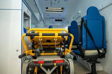 Arka kapılardan içeriye bakan temiz bir ambulans aracının geniş açılı görüntüsü. Parlak ışık altında katlanabilir sarı sedye mavi sağlık koltukları ve güvenlik ekipmanları.