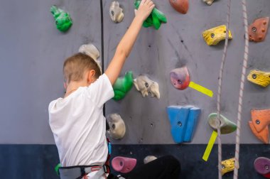 Beyaz tişört ve emniyet kemeri takmış okul çağındaki bir çocuk kapalı bir tırmanma duvarında eğitim görüyor. Spor dersi ya da hobisi sırasında profesyonel bir kayalık spor salonunda renkli durmaya çalışıyor.