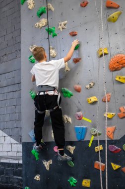 Beyaz tişört ve emniyet kemeri takmış okul çağındaki bir çocuk kapalı bir tırmanma duvarında eğitim görüyor. Spor dersi ya da hobisi sırasında profesyonel bir kayalık spor salonunda renkli durmaya çalışıyor.