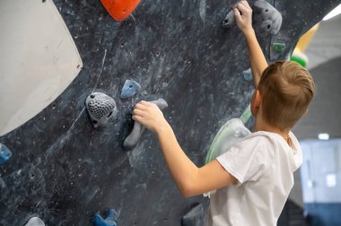 Beyaz tişörtlü bir öğrenci koyu dokulu bir tırmanma duvarı üzerinde çalışıyor. Spor dersi ya da aktif boş zamanlarında profesyonel bir kaya toplama merkezindeki gri ve mavi bölgelere uzanıyor.