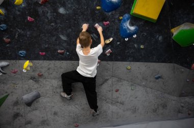 Beyaz tişörtlü ve siyah pantolonlu bir öğrenci karanlık bir duvarda antrenman yapıyor. Spor dersi sırasında ya da evde boş vakitlerinde profesyonel bir kaya atölyesinde tutunmaya çalışıyor.