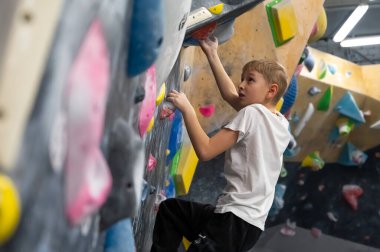 Beyaz tişörtlü mutlu bir okul çocuğu, eğitimi sırasında karanlık bir duvarda poz veriyor. Spor dersi ya da aktif hobisi sırasında profesyonel bir kaya atölyesinde kırmızı tutuş sergiliyor.