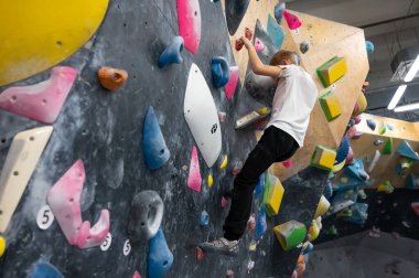 Beyaz tişörtlü ve siyah pantolonlu bir okul çocuğu koyu dokulu bir tırmanma duvarında eğitim görüyor. Spor dersi sırasında modern bir profesyonel kayalık merkezde renkli tutumlar sergiliyor.