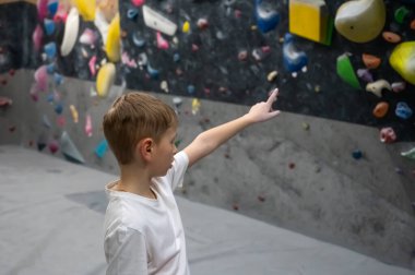 Beyaz tişörtlü bir okul çocuğu tırmanma yolunu koyu dokulu bir duvarda planlıyor. Spor dersi ya da aktif hobisi sırasında profesyonel bir kaya merkezindeki renkli duraklara işaret ediyor.