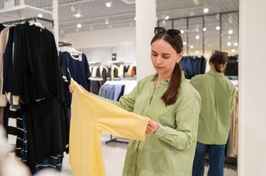 Perakende satış ve purchas için modern butik iç tasarımı olan bir giyim mağazasında askıdaki parlak sarı bir gömleğin kumaşını tutan ve kontrol eden kadın müşteri.