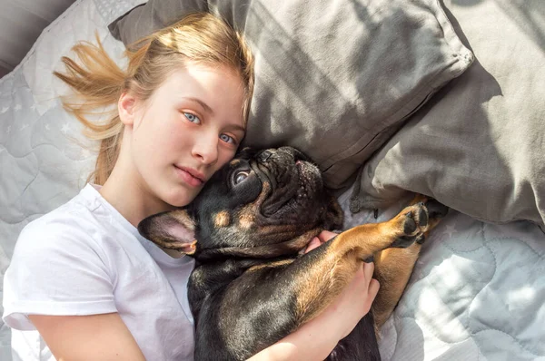 女の子はフランスのブルドッグ品種の彼女の犬と抱擁でベッドの上に横たわっている — ストック写真