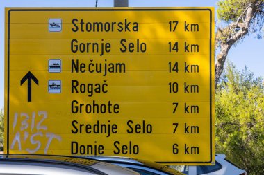 Parlak sarı bir yol tabelasında, Stomorska ve Grohote de dahil olmak üzere çok sayıda Hırvat ada kasabasına uzaklıklar sıralanıyor. Sahne, güneşli bir açık havada seyahat, seyrüsefer ve turistik yol tabelasını öneriyor..
