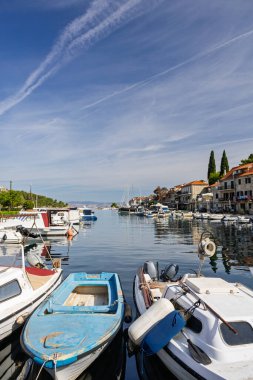 Güneşli bir sahil köyü boyunca demirlemiş yelkenli ve küçük teknelerle sakin bir liman sahnesi. Açık mavi gökyüzü, yumuşak su ve tarihi taş binalar huzur dolu deniz kenarlarını çağrıştırıyor..