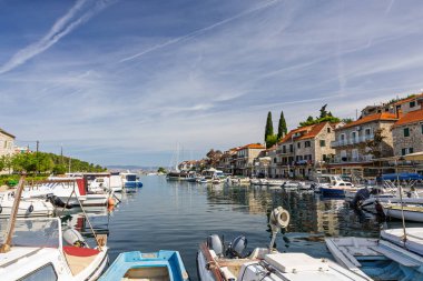 Güneşli bir sahil köyü boyunca demirlemiş yelkenli ve küçük teknelerle sakin bir liman sahnesi. Açık mavi gökyüzü, yumuşak su ve tarihi taş binalar huzur dolu deniz kenarlarını çağrıştırıyor..
