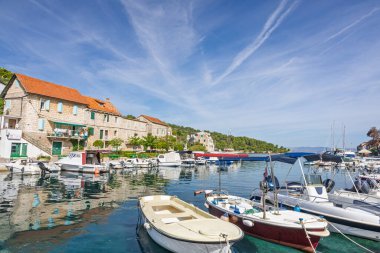 Güneşli bir sahil köyü boyunca demirlemiş yelkenli ve küçük teknelerle sakin bir liman sahnesi. Açık mavi gökyüzü, yumuşak su ve tarihi taş binalar huzur dolu deniz kenarlarını çağrıştırıyor..