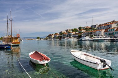 Küçük sandalların, demirli yelkenlilerin ve kırmızı kiremitli çatıları olan bir sahil köyünün olduğu sakin bir liman sahnesi. Sakin su, yukarıdaki damarları ve mavi gökyüzünü yansıtır, rahatlık ve sahil cazibesini aktarır..