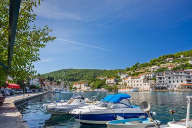 Kayık rıhtımına yanaşmış teknelerle dolu parlak bir liman manzarası, yamaçta pastel ve beyaz evler, ve huzurlu bir sahil köyü havası taşıyan açık mavi bir gökyüzü..
