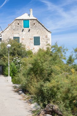 Turkuaz mavi panjurlu taştan bir ev, açık mavi bir gökyüzünün altında, çakıl yolunun ve yemyeşil bir yeşilliğin yanında oturur. Seyahat, mimari veya Akdeniz yaşam tarzı temaları için mükemmel.