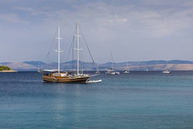 Sakin sahil limanında, güneşli bir gökyüzünün altında durgun mavi suyu noktalayan uzak tepeler ve diğer yatlarla küçük şişme bir teknenin yanında demirlemiş klasik bir ahşap yelkenli gemi var..
