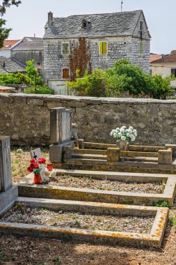 Güneşli bir kırsal kesimde alçak duvarın arkasında yıpranmış taş bir ev, ön planda çiçeklerle süslenmiş küçük bir mezarlık görülüyor. Bereketli yeşillik, huzurlu ve nostaljik bir manzarayı çerçeveler.