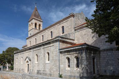 Ayırt edici bir çan kulesinin bulunduğu eski taş kilise, yemyeşil bir çan kulesiyle çevrili ve berrak, mavi bir gökyüzüne karşı güneş ışığıyla yıkanmış, dingin ve ruhani bir atmosferi resmediyor..
