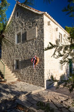 NECUJAM, CROATIA, 09.01.2025: Bir taş bina duvarında kırmızı, beyaz ve mavi kurdeleli bir çelenk ile bir hatıra plaketi barındırır. Güneşli adımlar ve derin mavi bir gökyüzü sakin tarihi bir sokak sahnesini canlandırıyor..