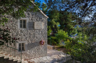 NECUJAM, CROATIA, 09.01.2025: Bir taş bina duvarında kırmızı, beyaz ve mavi kurdeleli bir çelenk ile bir hatıra plaketi barındırır. Güneşli adımlar ve derin mavi bir gökyüzü sakin tarihi bir sokak sahnesini canlandırıyor..