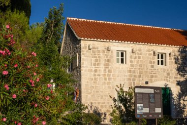 NECUJAM, CROATIA, 09.01.2025: Bir taş bina duvarında kırmızı, beyaz ve mavi kurdeleli bir çelenk ile bir hatıra plaketi barındırır. Güneşli adımlar ve derin mavi bir gökyüzü sakin tarihi bir sokak sahnesini canlandırıyor..